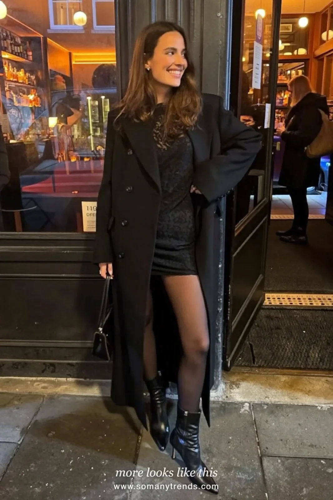 Woman in a black lace mini dress under a long black coat with sheer tights and heeled boots outside a bar, outfit 22