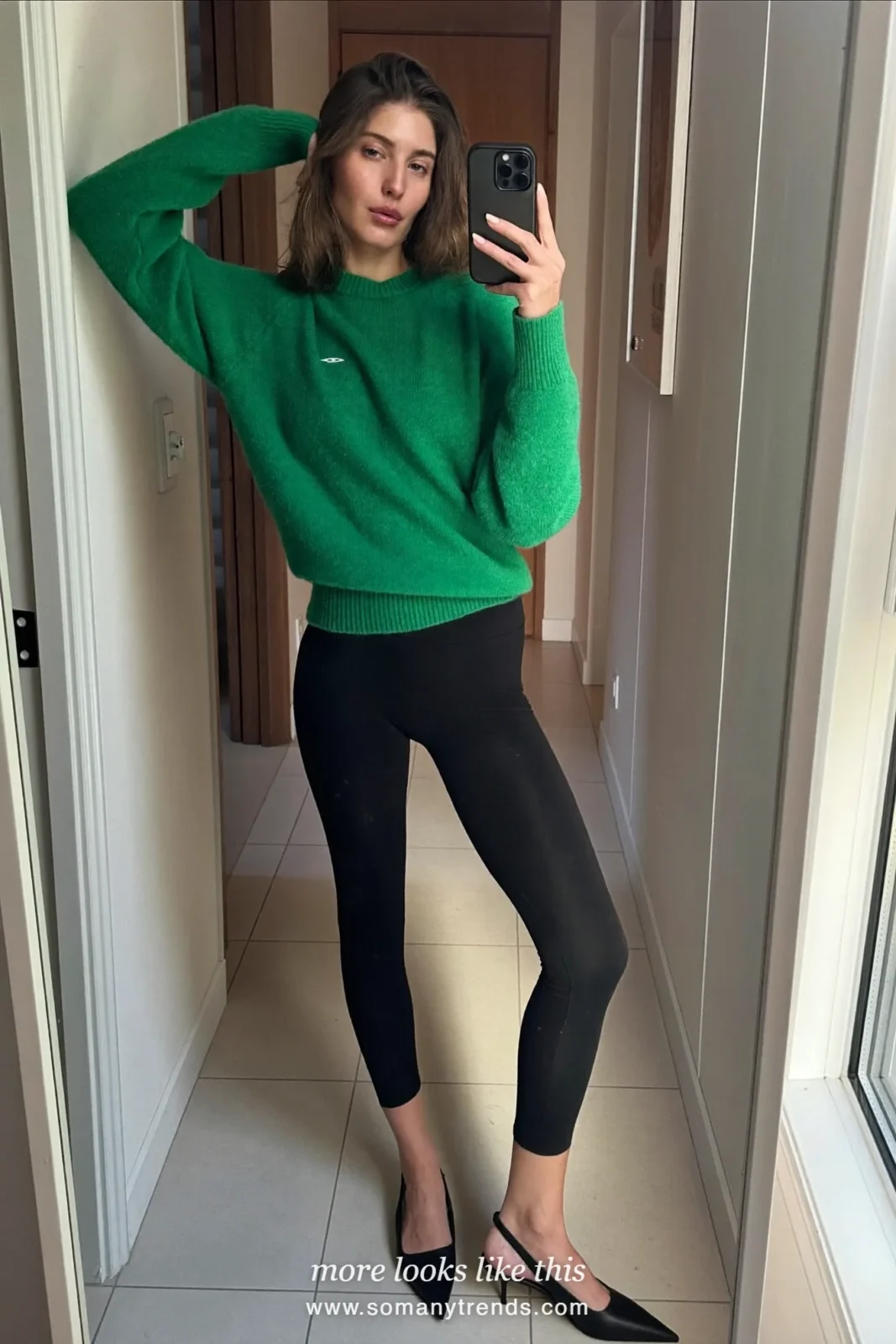 Woman in an emerald fine-knit sweater, black leggings and black slingback heels taking a mirror selfie at home, look 4