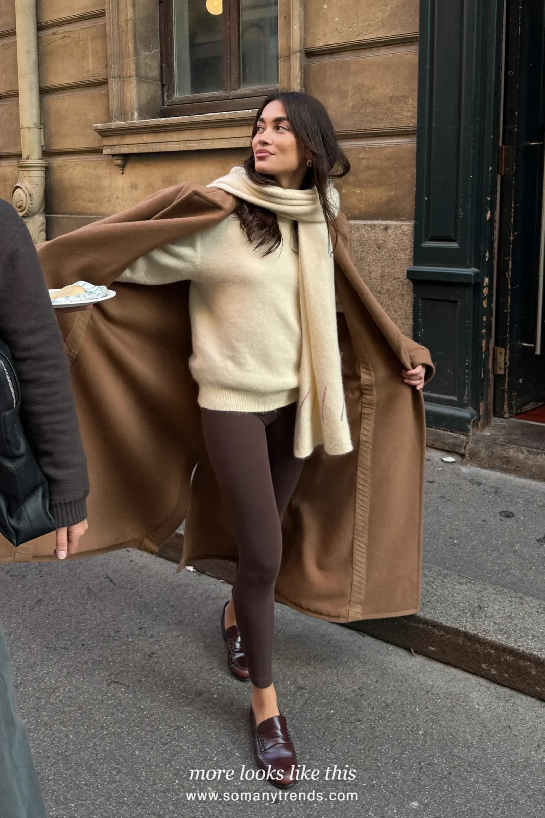 Woman in a long camel coat, chocolate brown leggings, cream scarf and brown loafers, look 6