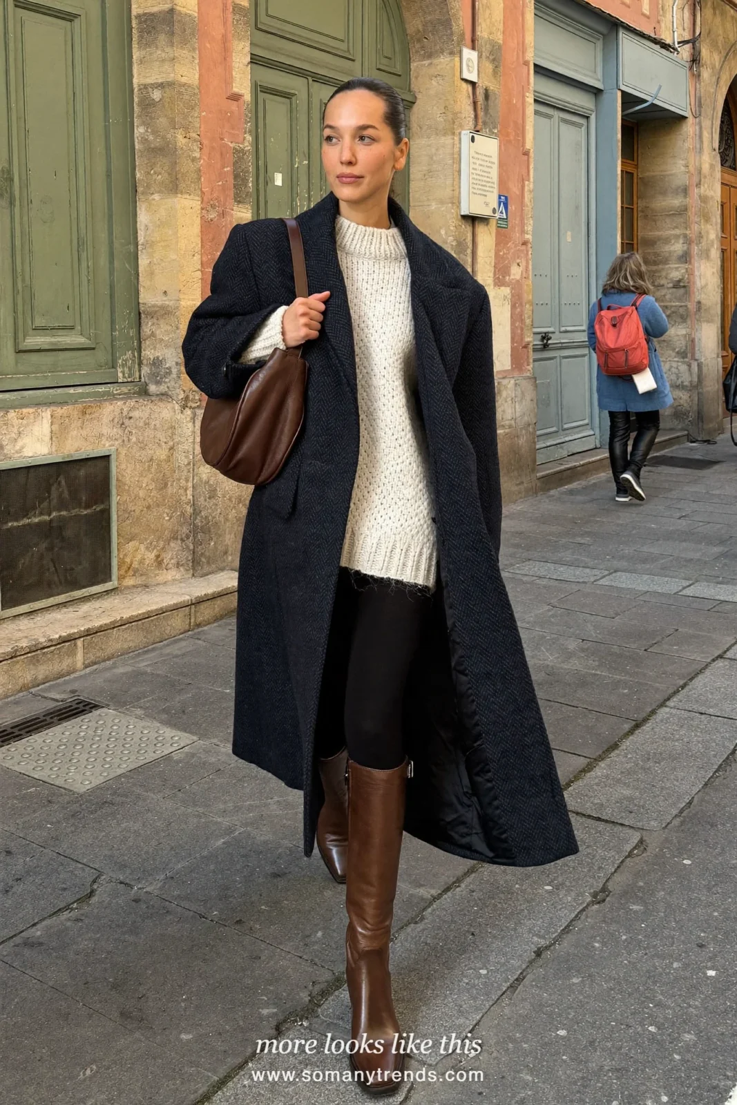 Woman in a long navy coat, cream sweater, black leggings and brown leather boots, look 22