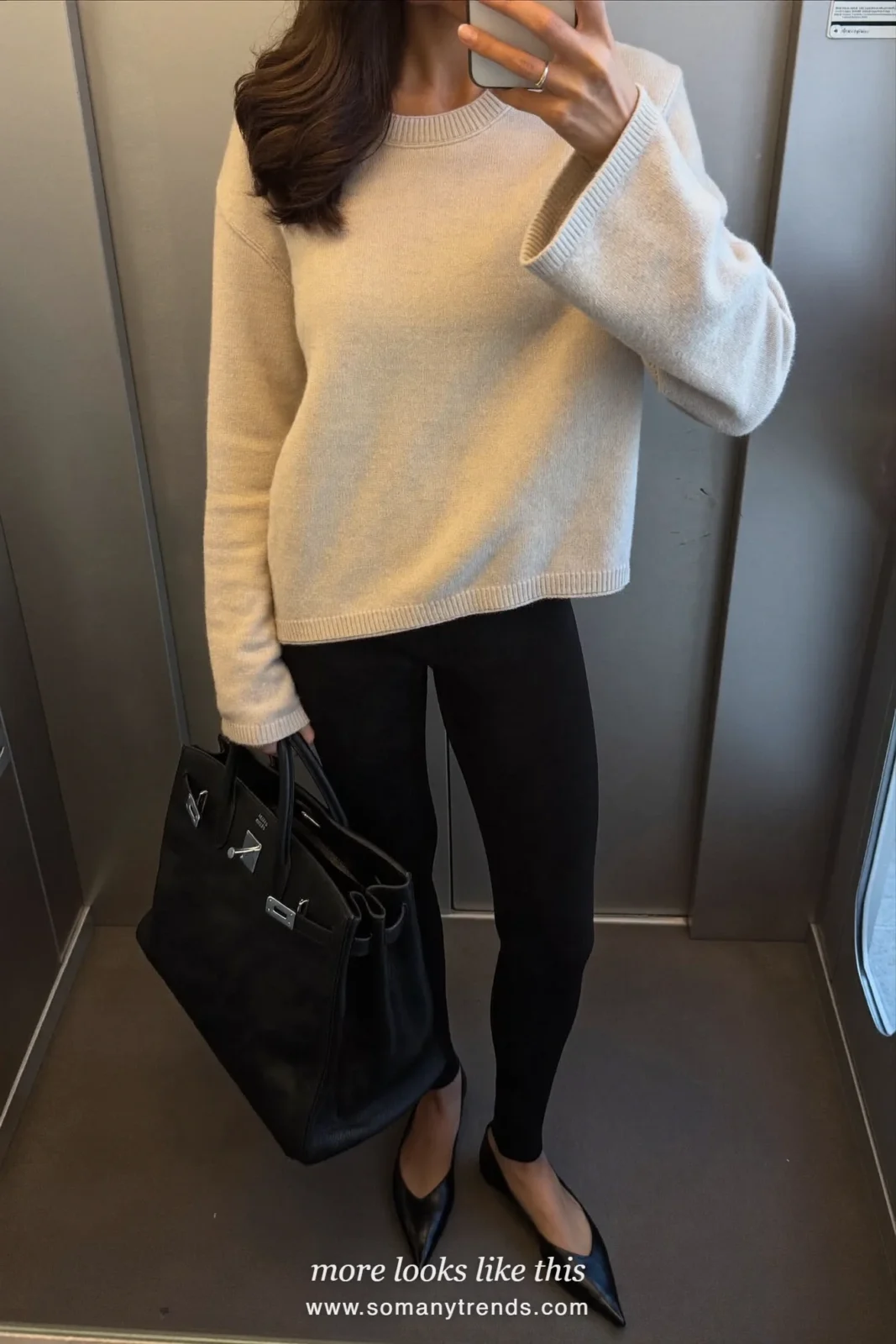 Woman in a black wool coat, cream sweater, black leggings and pointed flats taking a mirror selfie, look 28