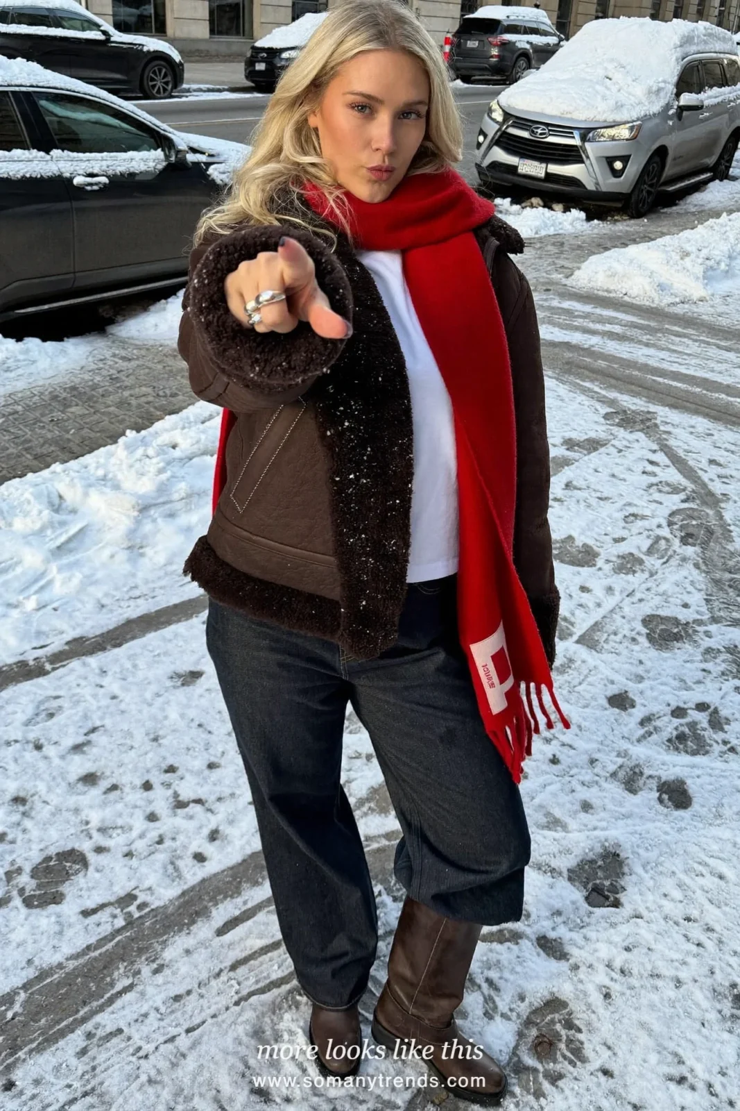 Shearling aviator jacket with red scarf, dark jeans and tall brown boots on a snowy street, outfit 14