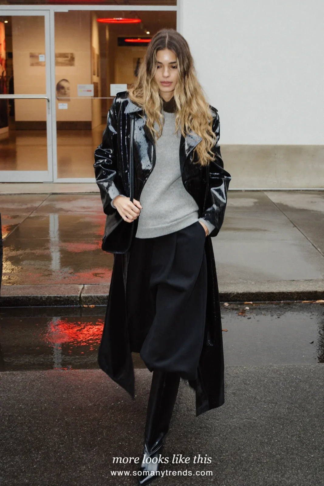 Woman in black patent belted long trench coat, gray knit, black pants and black knee boots, look 5