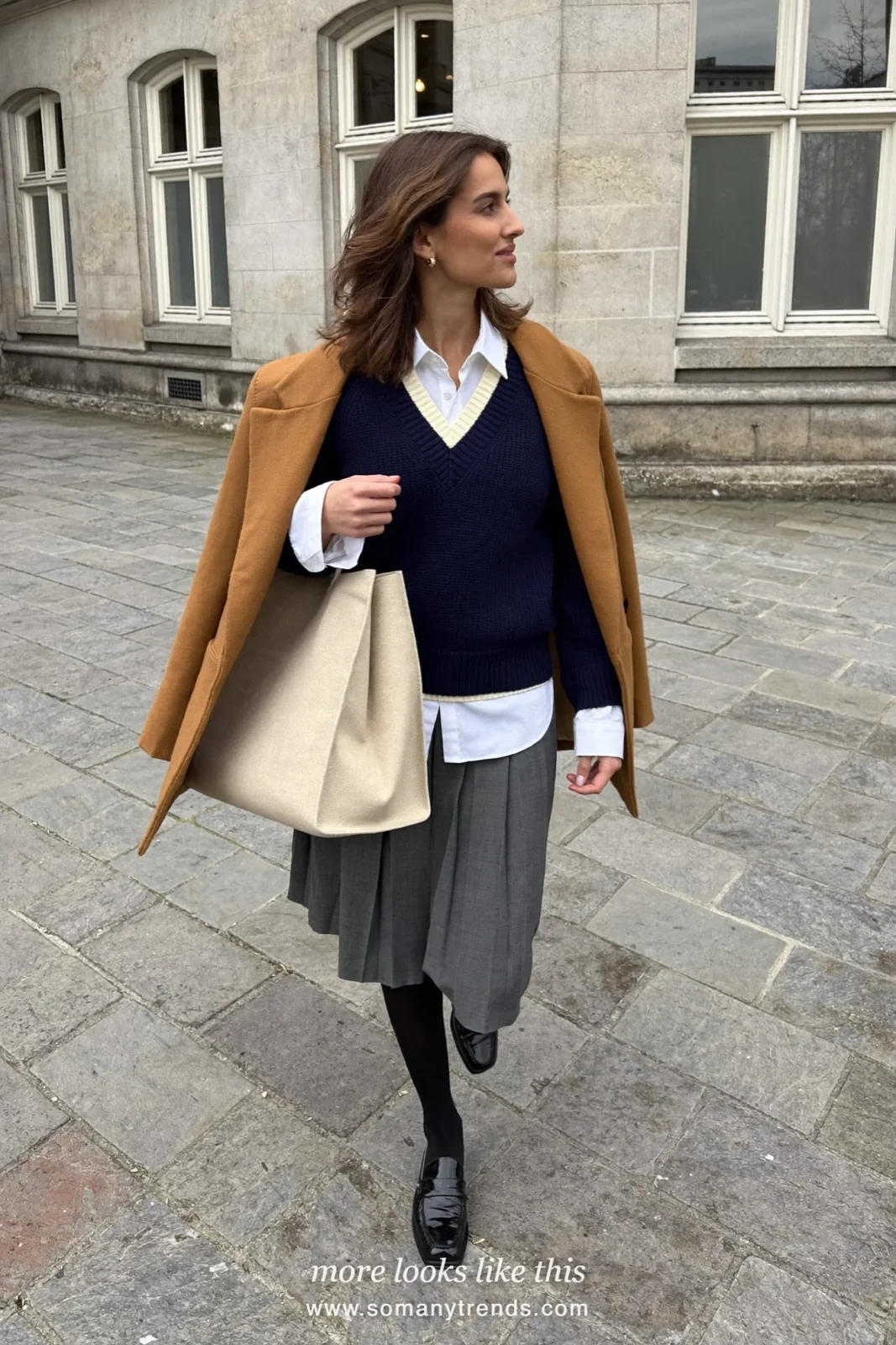 Woman in navy V-neck knit with cream trim, white collared shirt, gray pleated midi skirt, black tights and loafers, look 6