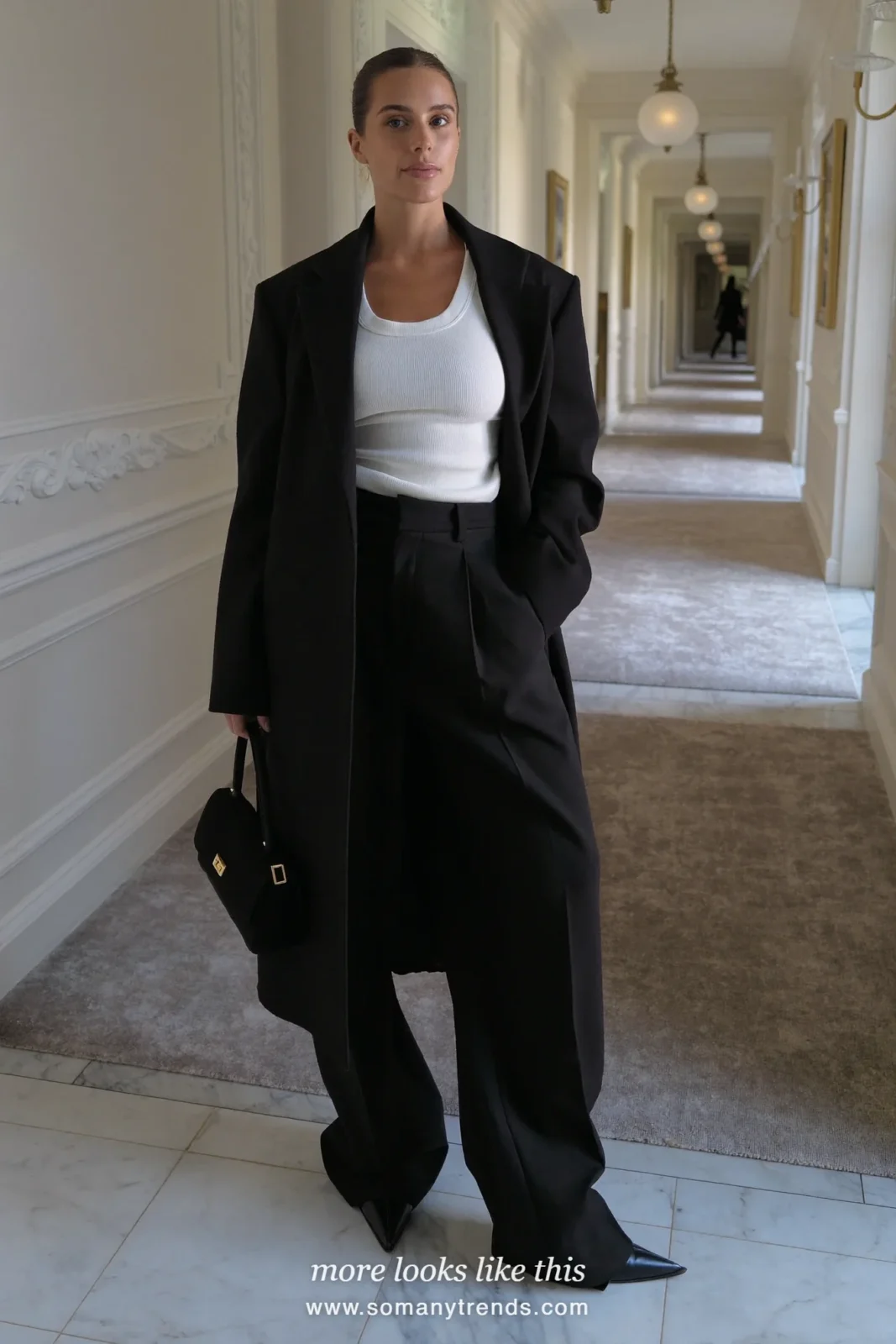 Woman in a long black coat over a white tank and wide black pants in a hotel corridor, outfit 10