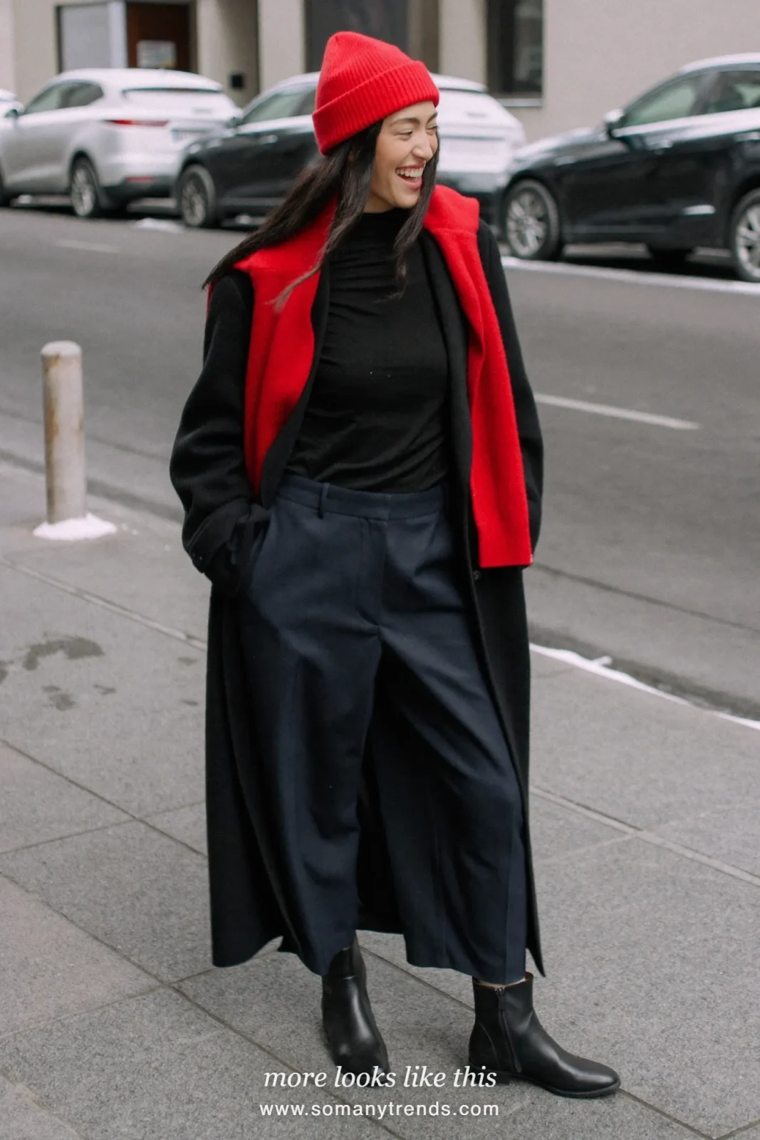 Long black wool coat with bright red beanie, red knit over the shoulders, dark pants and black boots on a snowy city street, outfit 3