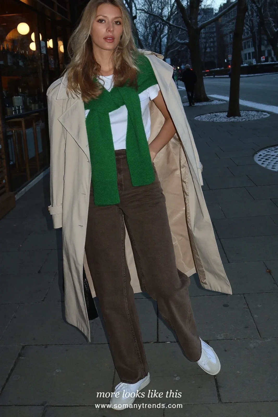 Woman in chocolate brown wide-leg jeans, trench coat, green sweater over the shoulders and white sneakers, outfit 3