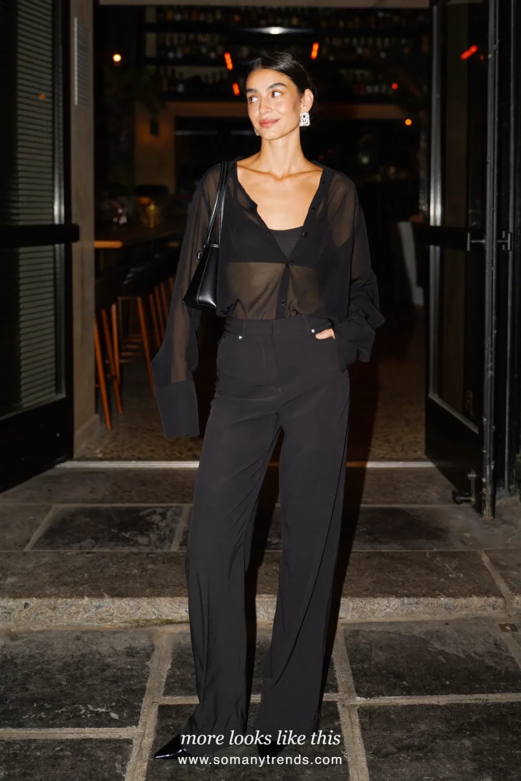 Woman in a sheer black blouse, camisole, wide black pants and black heels at a bar entrance, outfit 27