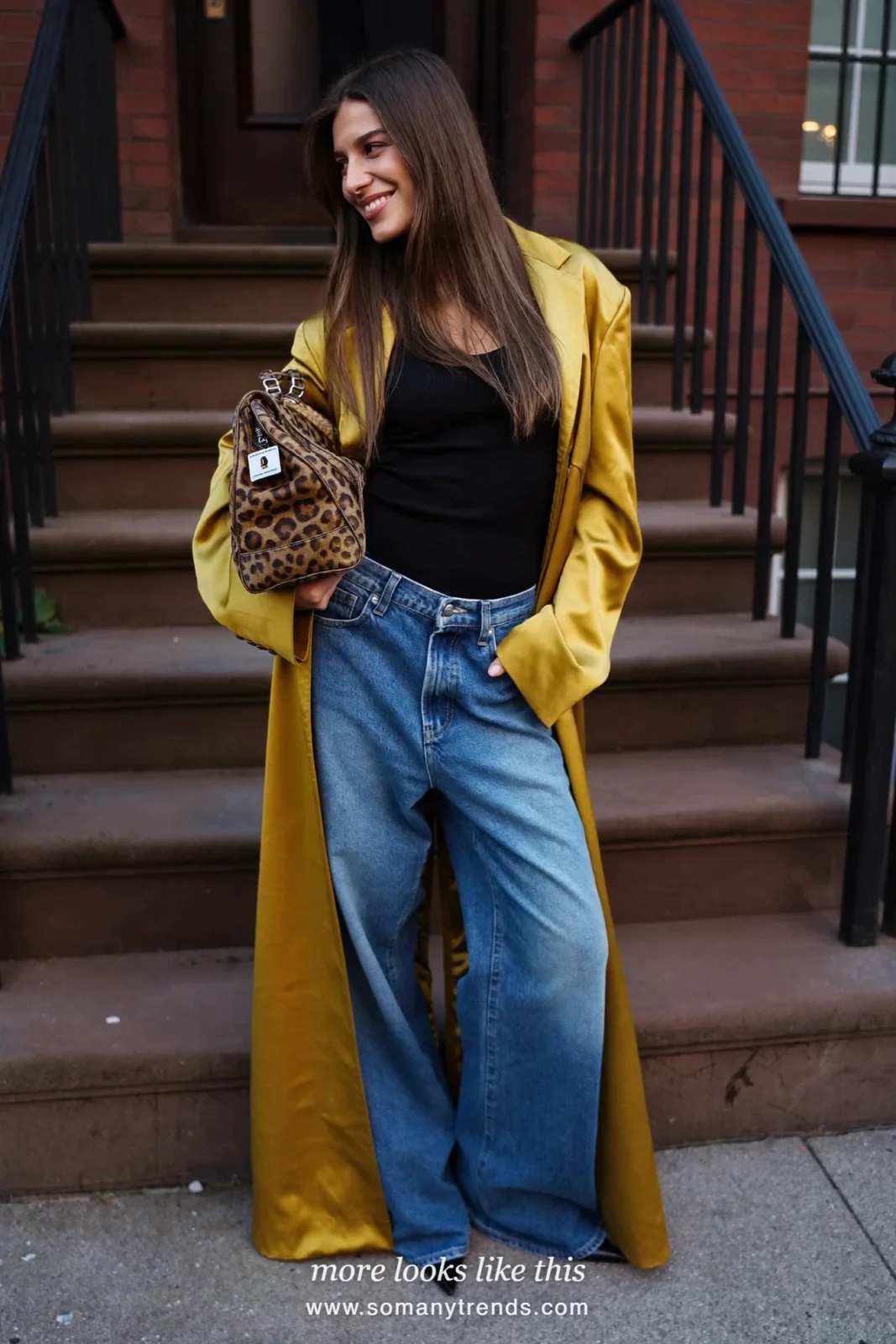 Woman on a brownstone stoop in a long mustard-gold satin coat, black top, wide-leg jeans, black pointed boots and a leopard clutch, outfit 1