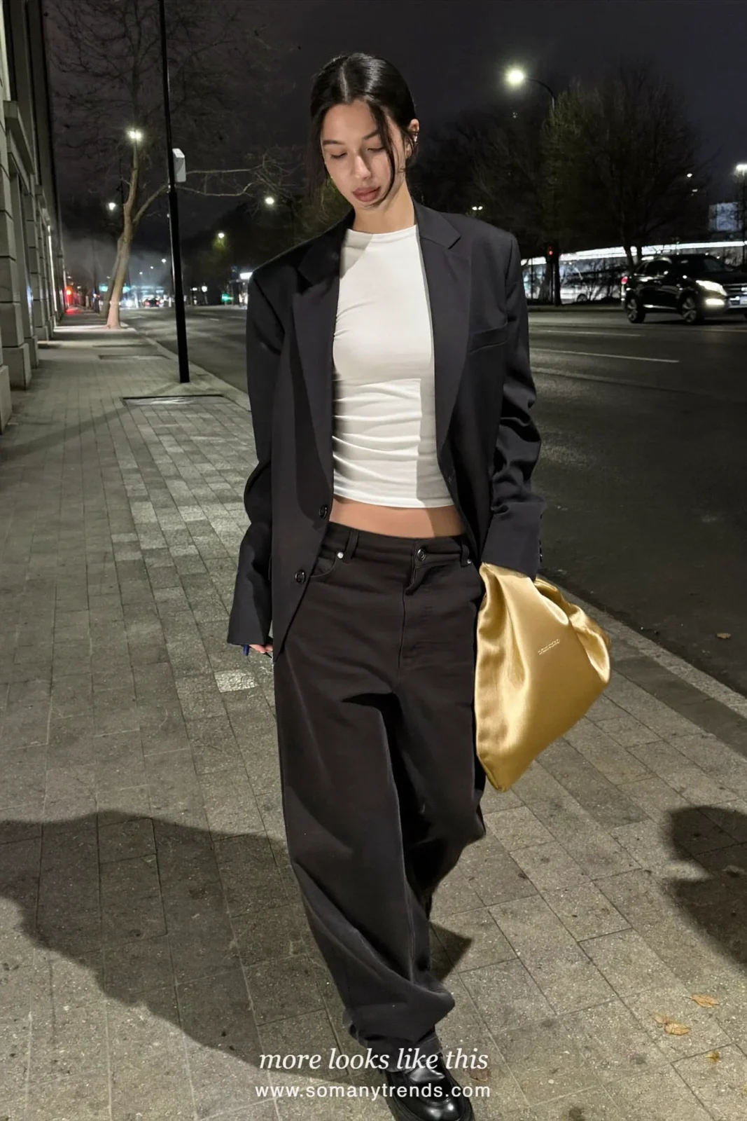 Woman in a black blazer, black wide-leg pants, black loafers and a gold satin pouch bag on a city sidewalk at night, outfit 3