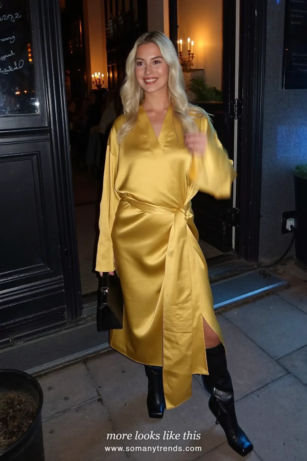 Woman in a gold satin belted wrap dress and black knee-high boots in a candle-lit bar, outfit 6