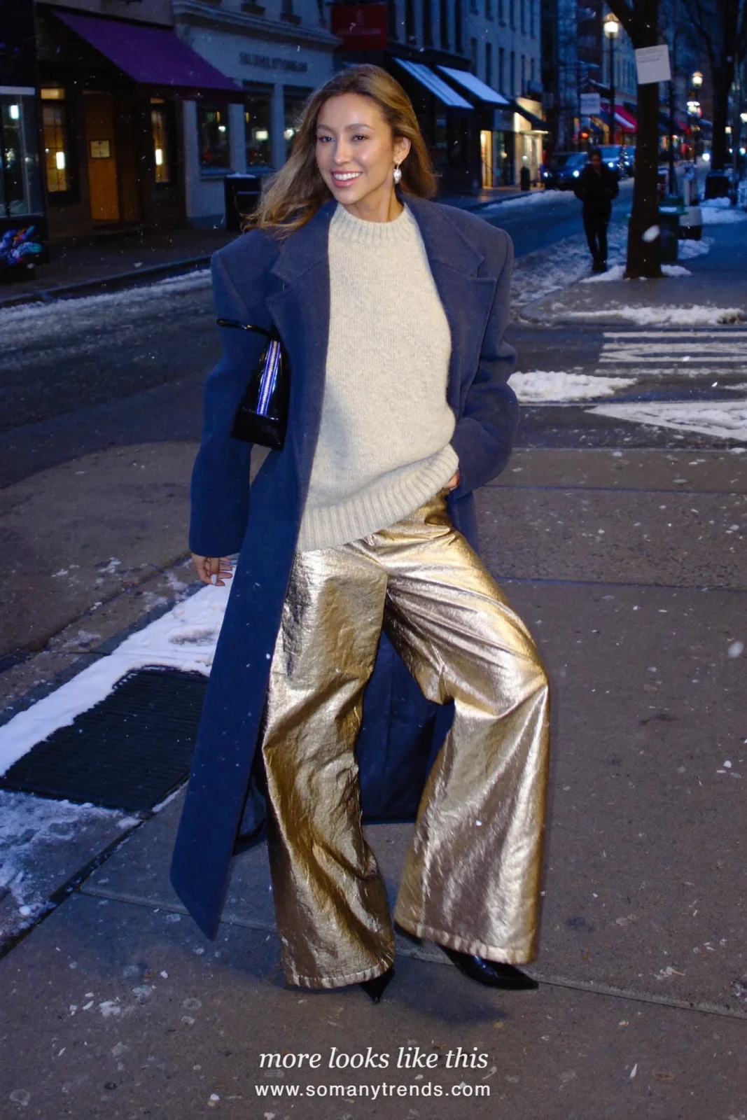 Woman in a long navy wool coat, cream knit, gold metallic wide-leg pants and black boots on a snowy city street, outfit 18
