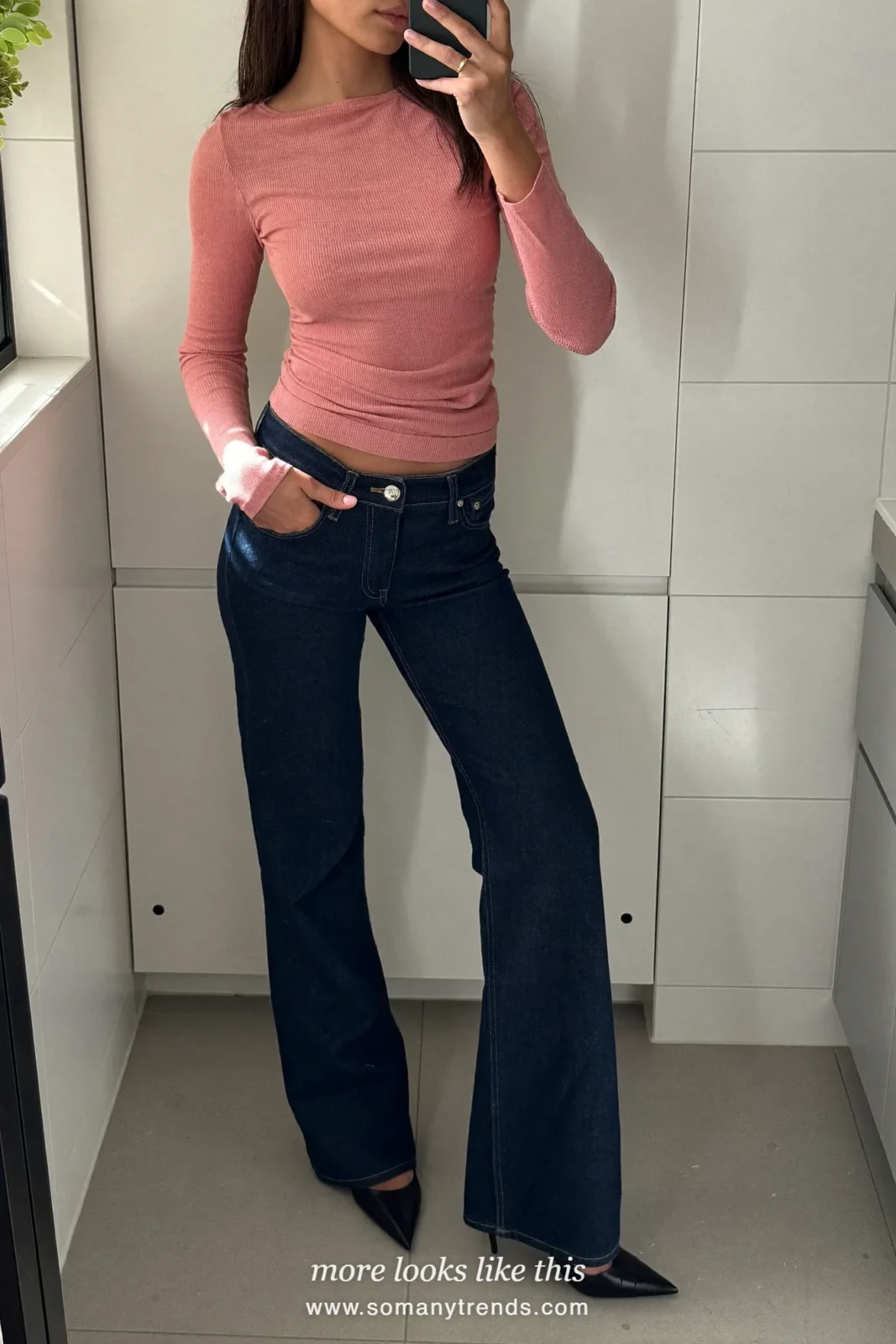 Woman in a rose knit top, dark indigo flare jeans and pointed pumps taking a mirror selfie in a tiled bathroom, outfit 4