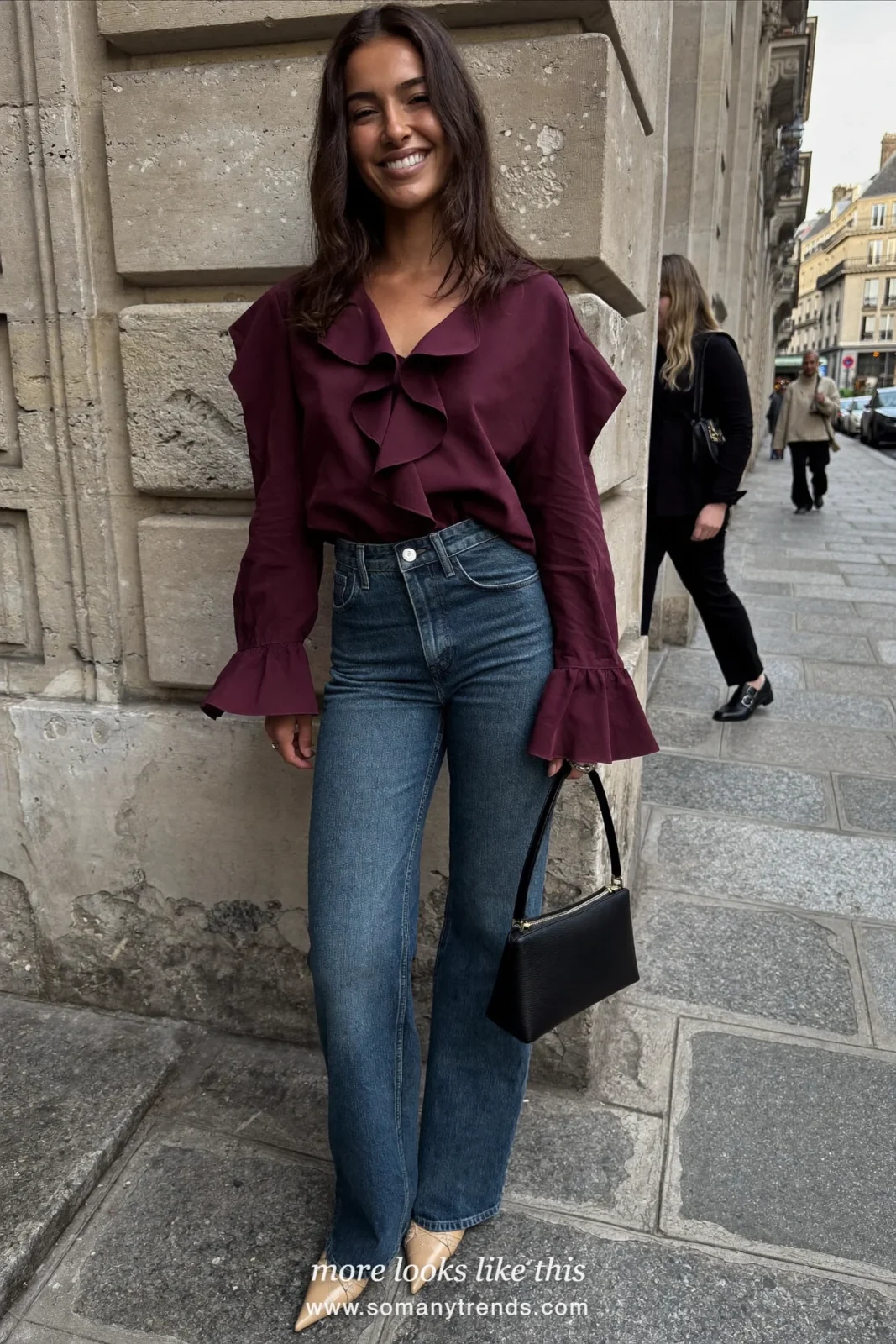 Woman in a burgundy ruffle-sleeve blouse, mid-blue flare jeans and beige heels near a stone wall, outfit 7