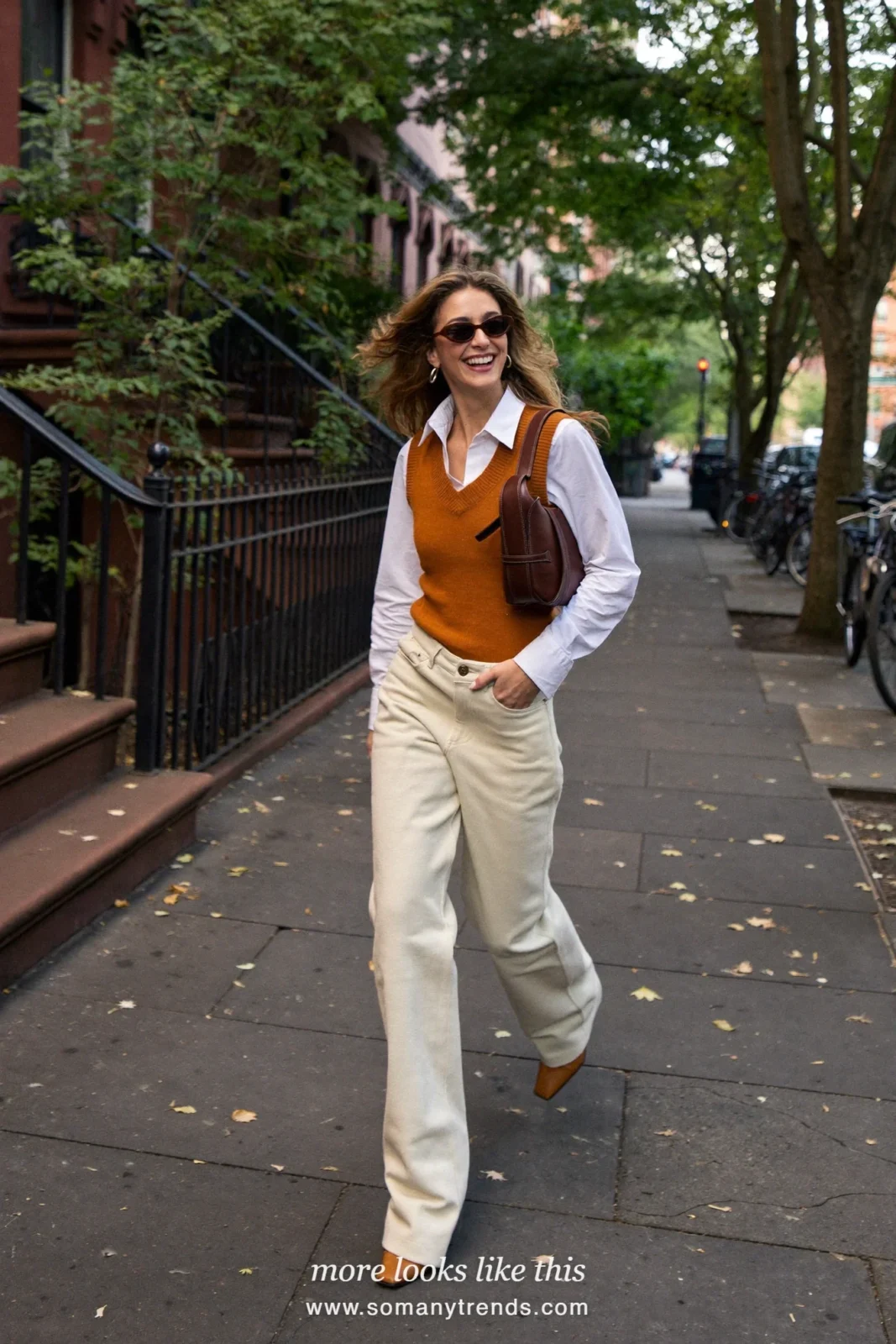 Woman in a rust knit vest, white shirt, cream flare pants and tan heeled boots on a city street, outfit 14