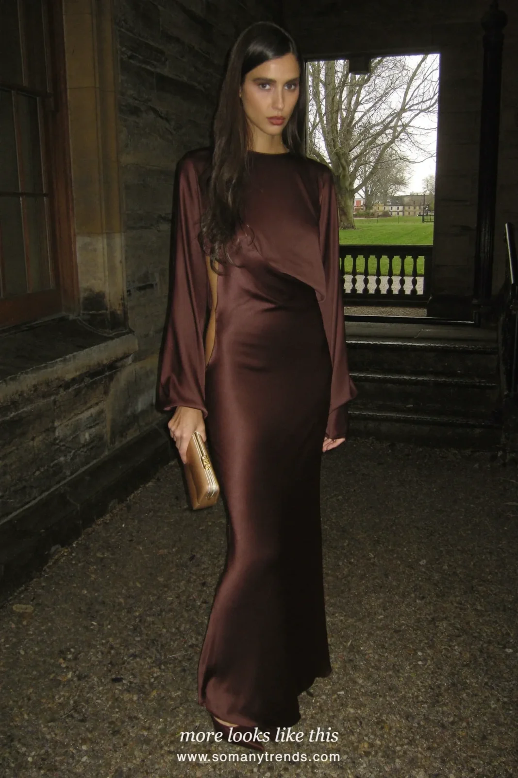Woman in a chocolate brown bias-cut satin gown with gold earrings and brown heels at a country manor entrance, outfit 11