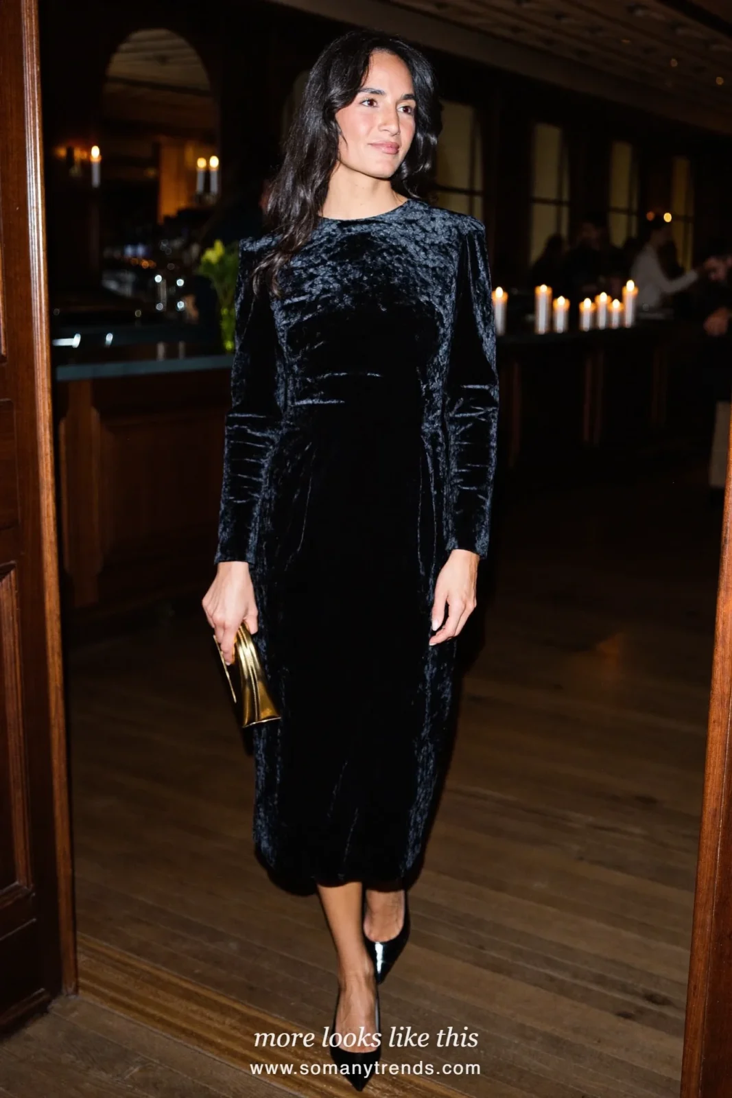 Woman in a black velvet long-sleeve column dress with a draped back and black pumps in a candlelit bar with dark wood, outfit 14