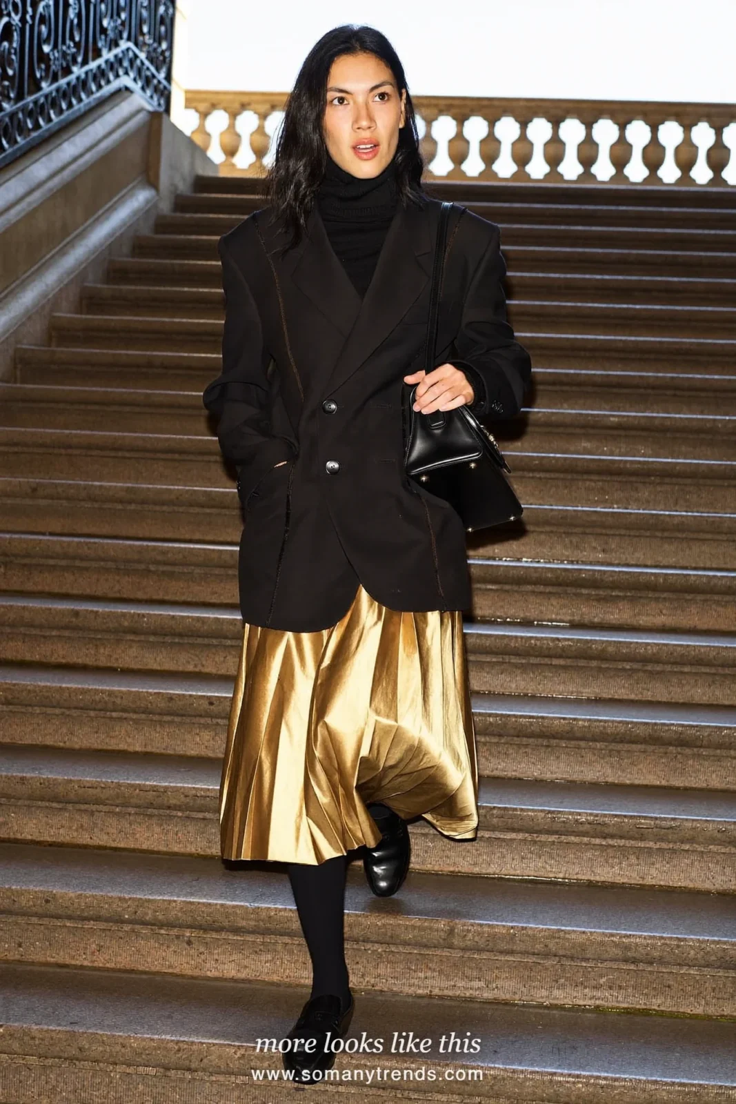 Woman in a gold pleated midi skirt, black turtleneck, oversize black blazer and black loafers on a staircase, outfit 14