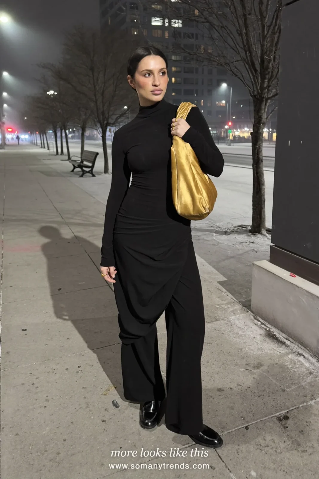 Woman in an all-black outfit with a black turtleneck, wide pants, black loafers and a gold satin bag on a sidewalk at night, outfit 25