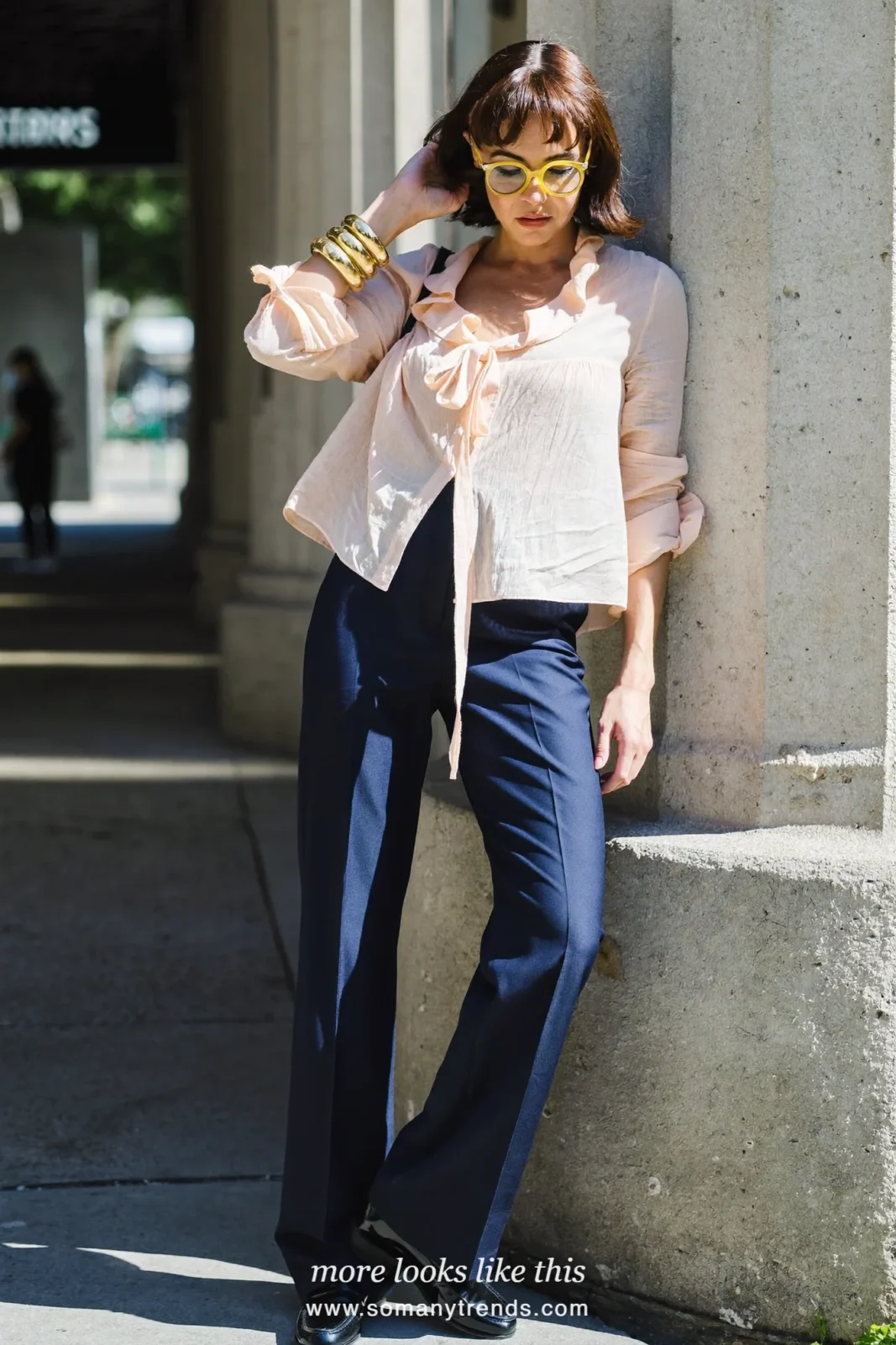 Woman in a pale pink ruffled tie-neck blouse, dark navy blue wide-leg pants, black mini bag and loafers, outfit 4