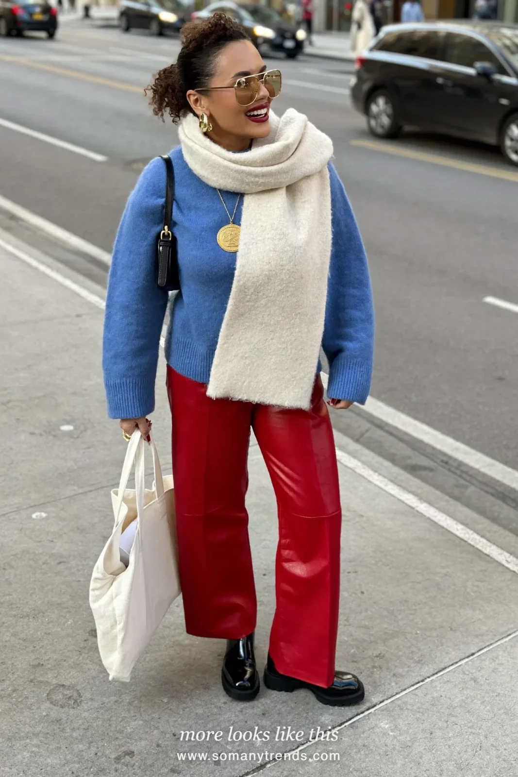 Woman in a cobalt blue fine knit sweater, cream scarf, red soft matte leather straight pants and black ankle boots, look 16