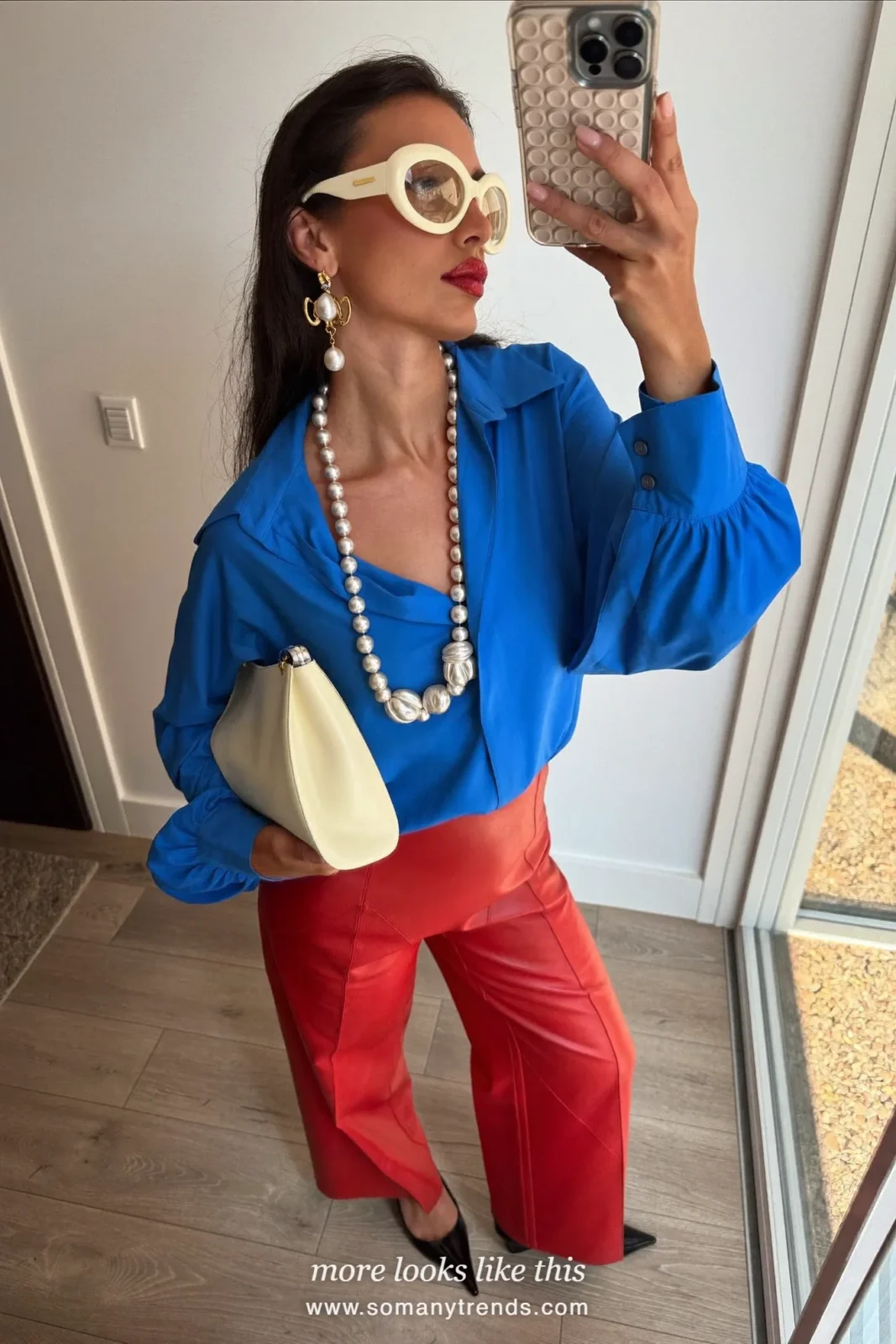 Woman in a cobalt blue silk blouse, red soft matte leather straight pants, black pointed pumps and a cream clutch, look 24