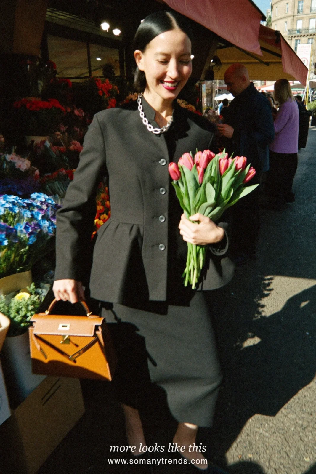 Petite woman in a black structured peplum jacket with mandarin collar, black column midi skirt, black pointed pumps and a cognac Kelly bag, outfit 1