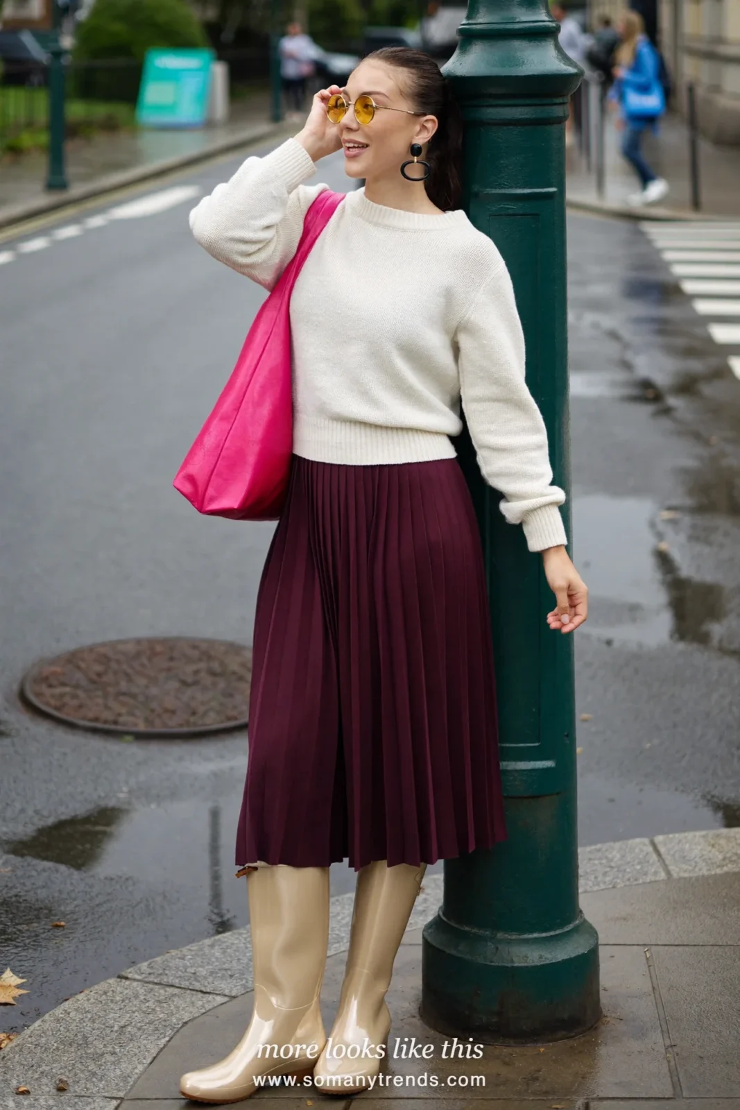 Woman in light metallic bronze tall rubber rain boots, plum midi skirt, cream knit sweater and hot pink tote on a damp street, outfit 1