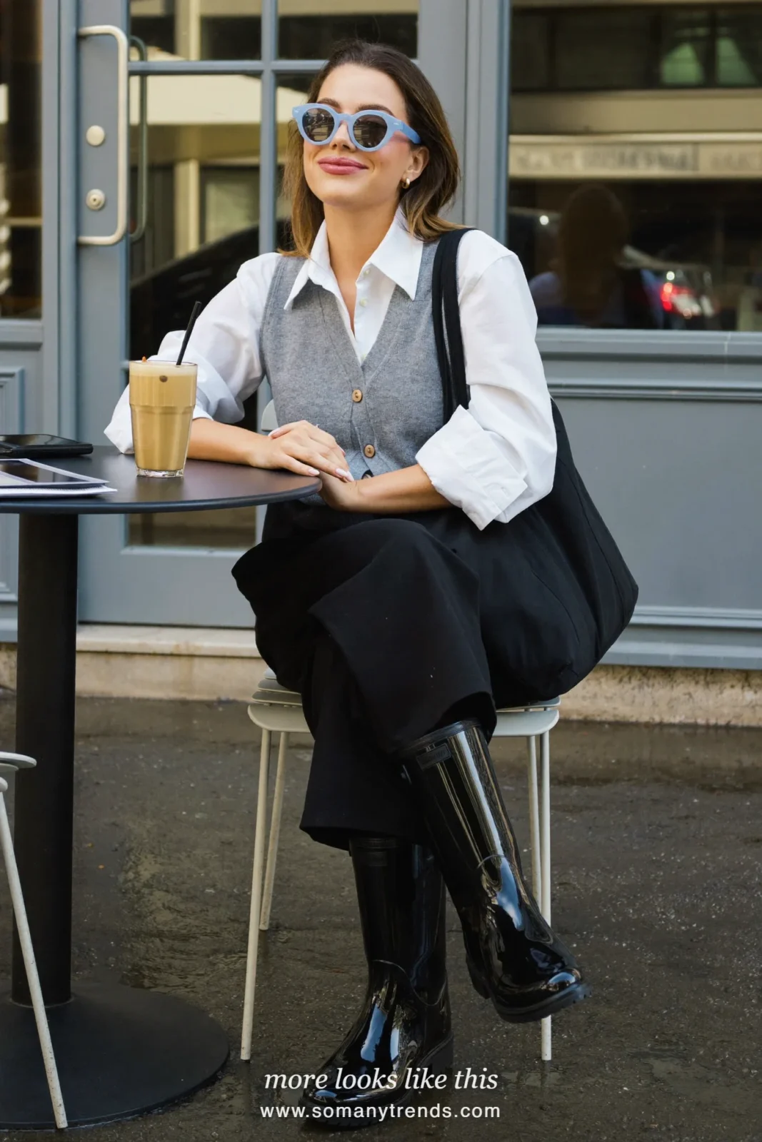 Woman in glossy black rain boots under wide-leg black pants, white shirt and gray knit vest outside an office, outfit 11