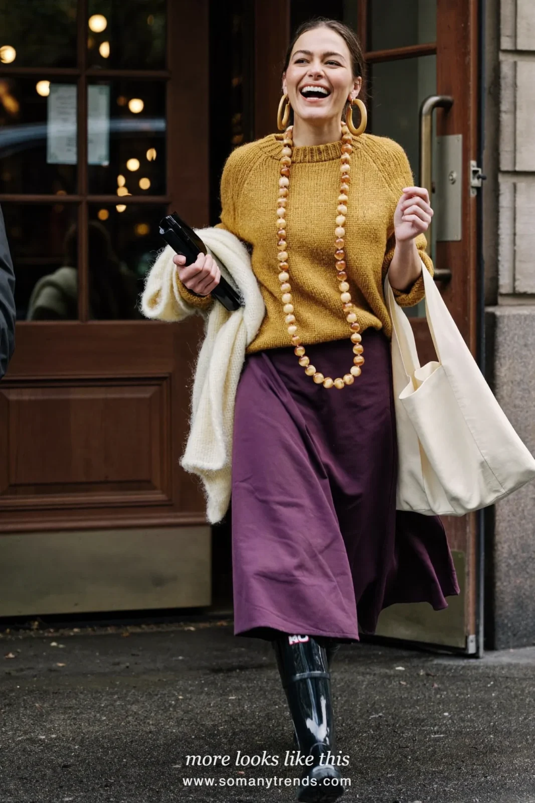 Woman in glossy black short rain boots, plum midi skirt and mustard knit with cream scarf, outfit 26