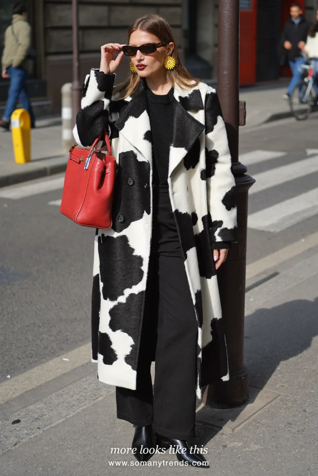 Woman in a long black and white cow print coat over a black knit, black wide pants and black boots with a cherry red bag, outfit 20