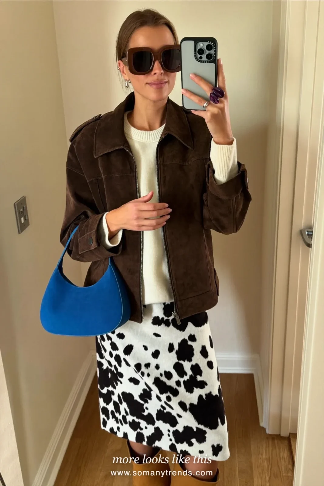 Woman in a black and white cow print midi skirt with a chocolate brown suede jacket, cream knit, tan boots and a cobalt blue bag, outfit 24