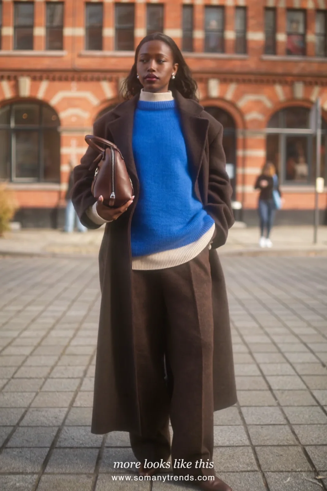 Woman in a long chocolate brown coat with a cobalt blue knit over her shoulders, cream knit, brown pants and brown boots, outfit 13