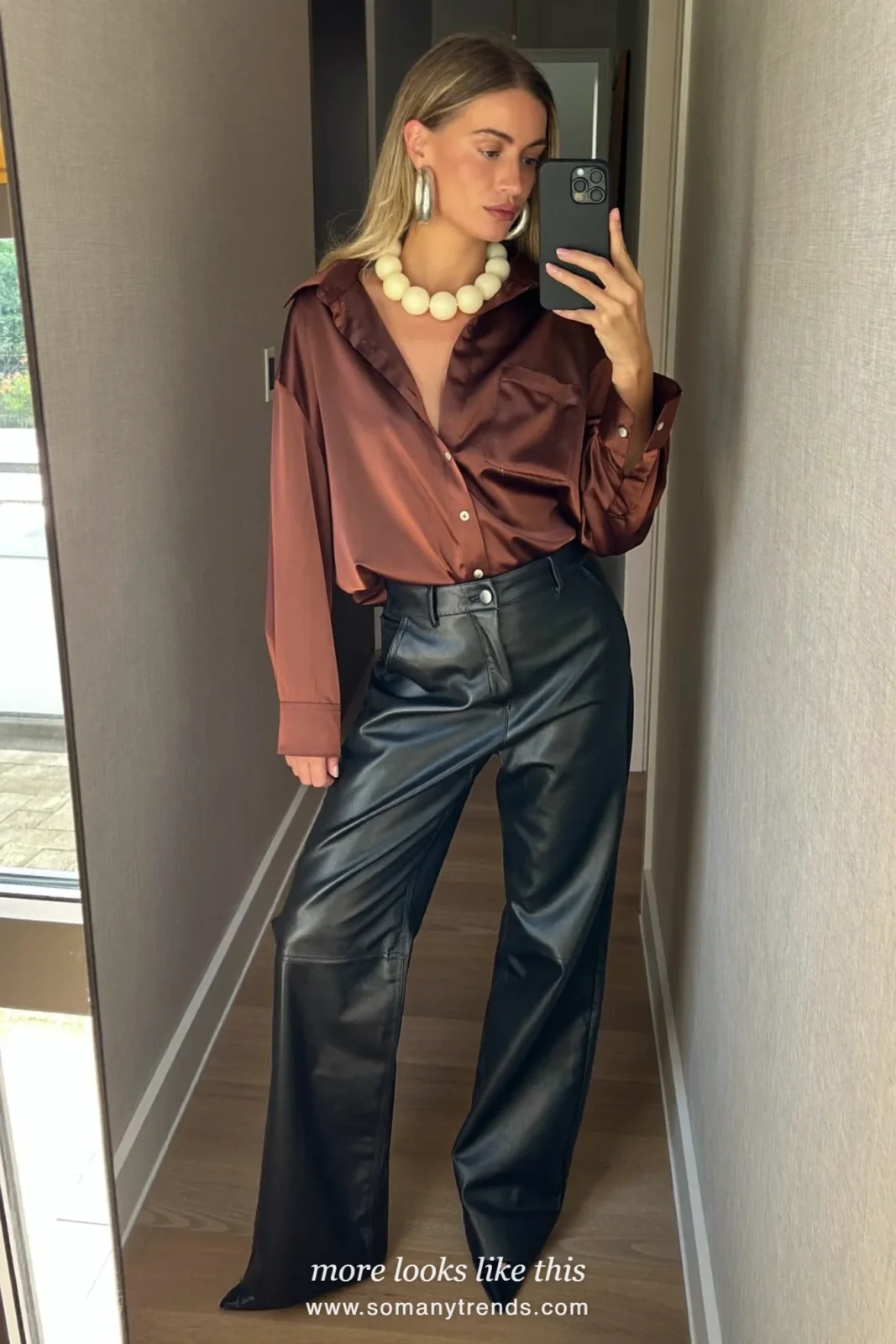 Woman in a chocolate brown satin shirt, black faux leather pants, black pointed heels and a cobalt blue bag, look 15