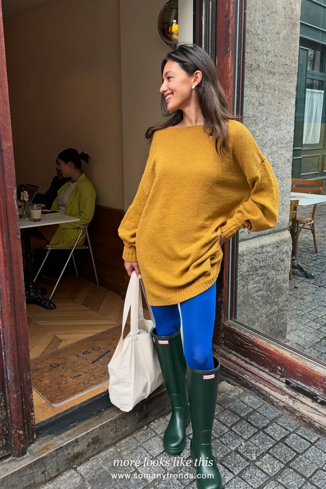 Woman in dark green glossy tall rain boots, mustard knit mini dress and cobalt opaque tights outdoors on wet pavement, outfit 2
