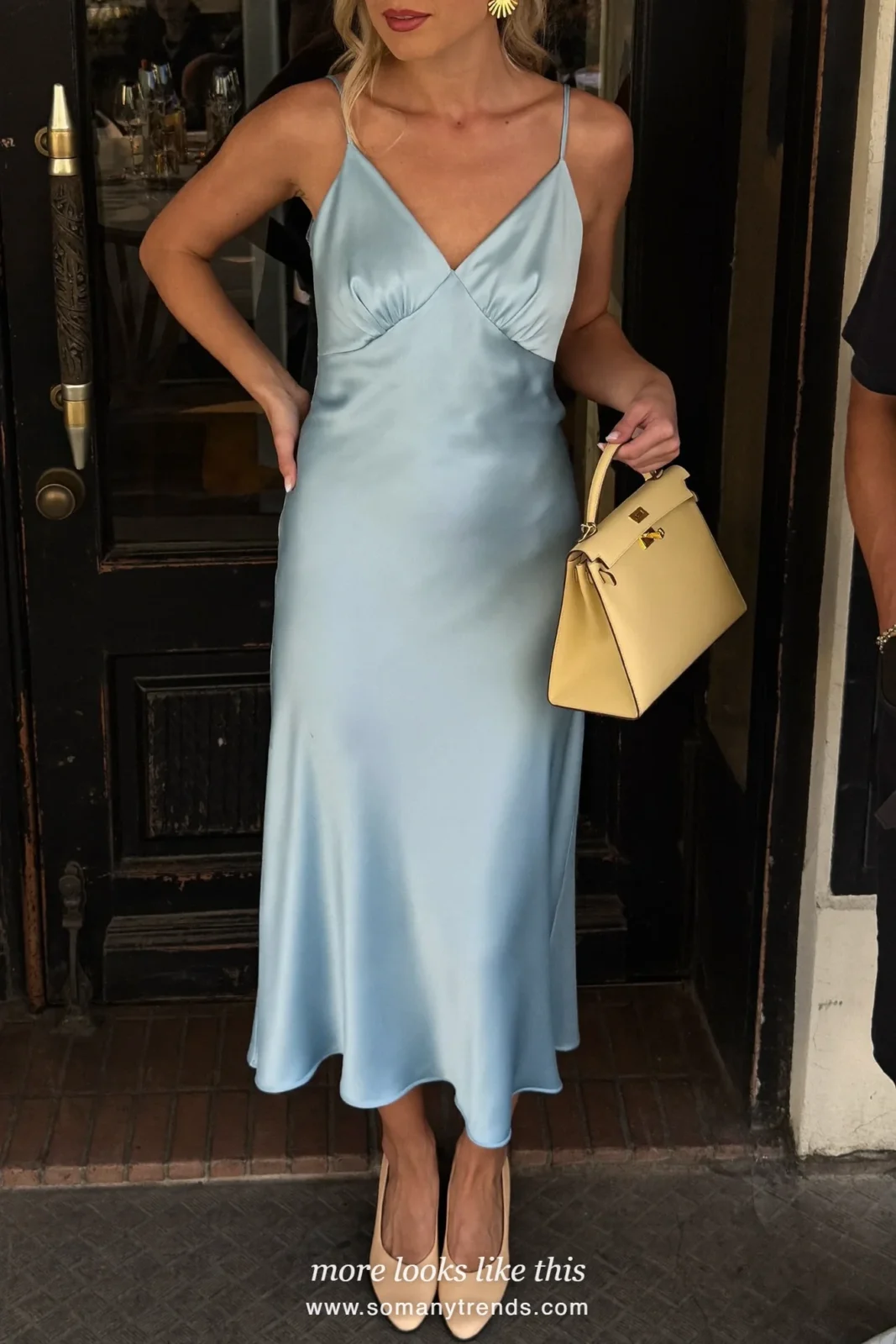 Woman in an ice blue satin slip midi dress, butter yellow top-handle bag, silver jewelry and nude pumps at a venue entrance, outfit 13