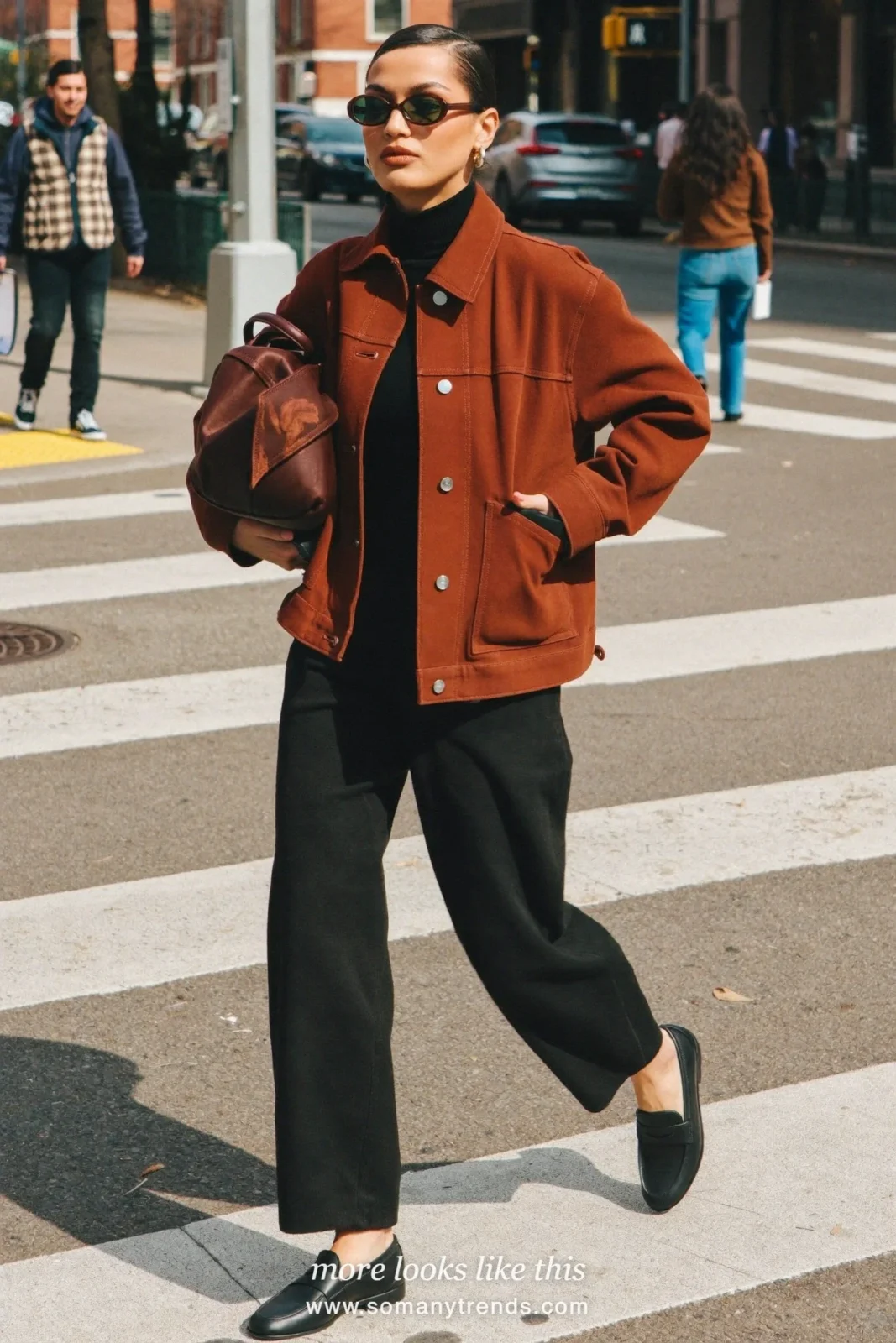 Woman in a rust brown work jacket, black turtleneck, black wide-leg pants, black loafers and an olive green tote bag, look 2