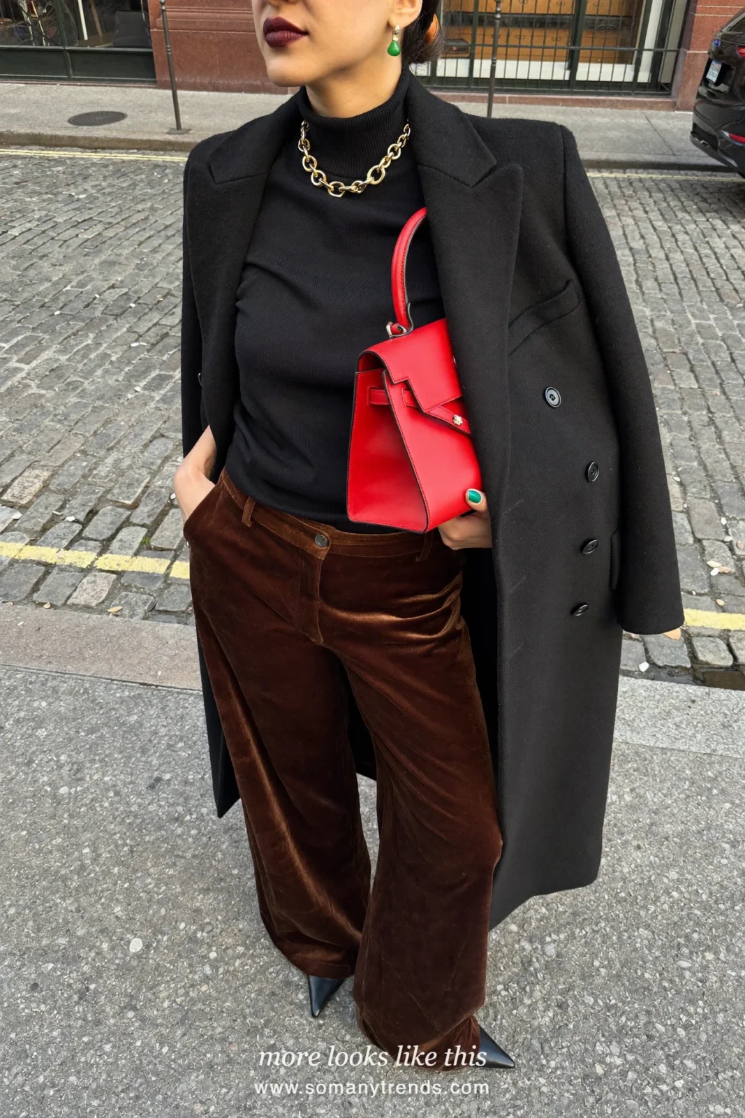 Woman in brown velvet wide-leg pants, a black fine knit top, a black coat and black pointed pumps with a cherry red bag, look 17