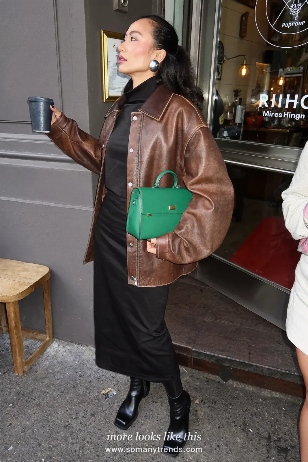 Woman in a brown leather jacket over a black midi dress with black tights, black boots and an emerald green bag, look 22