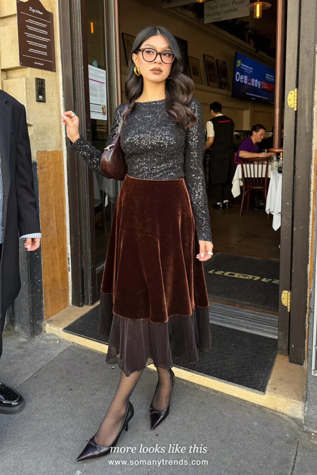 Woman in a black sequin long-sleeve top, brown velvet midi skirt, sheer black tights, black pointed pumps and a cherry red clutch, look 25