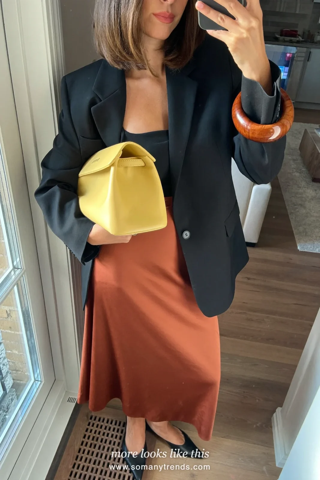 Woman in a black tuxedo blazer, black camisole, rust brown satin midi skirt, black pointed heels and a butter yellow clutch, look 27