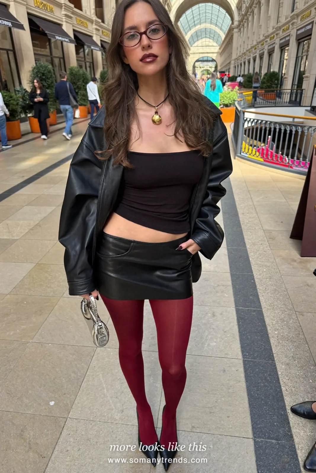 Woman in a black boxy jacket, black mini skirt, opaque burgundy tights, black pumps and a silver pouch, outfit 1
